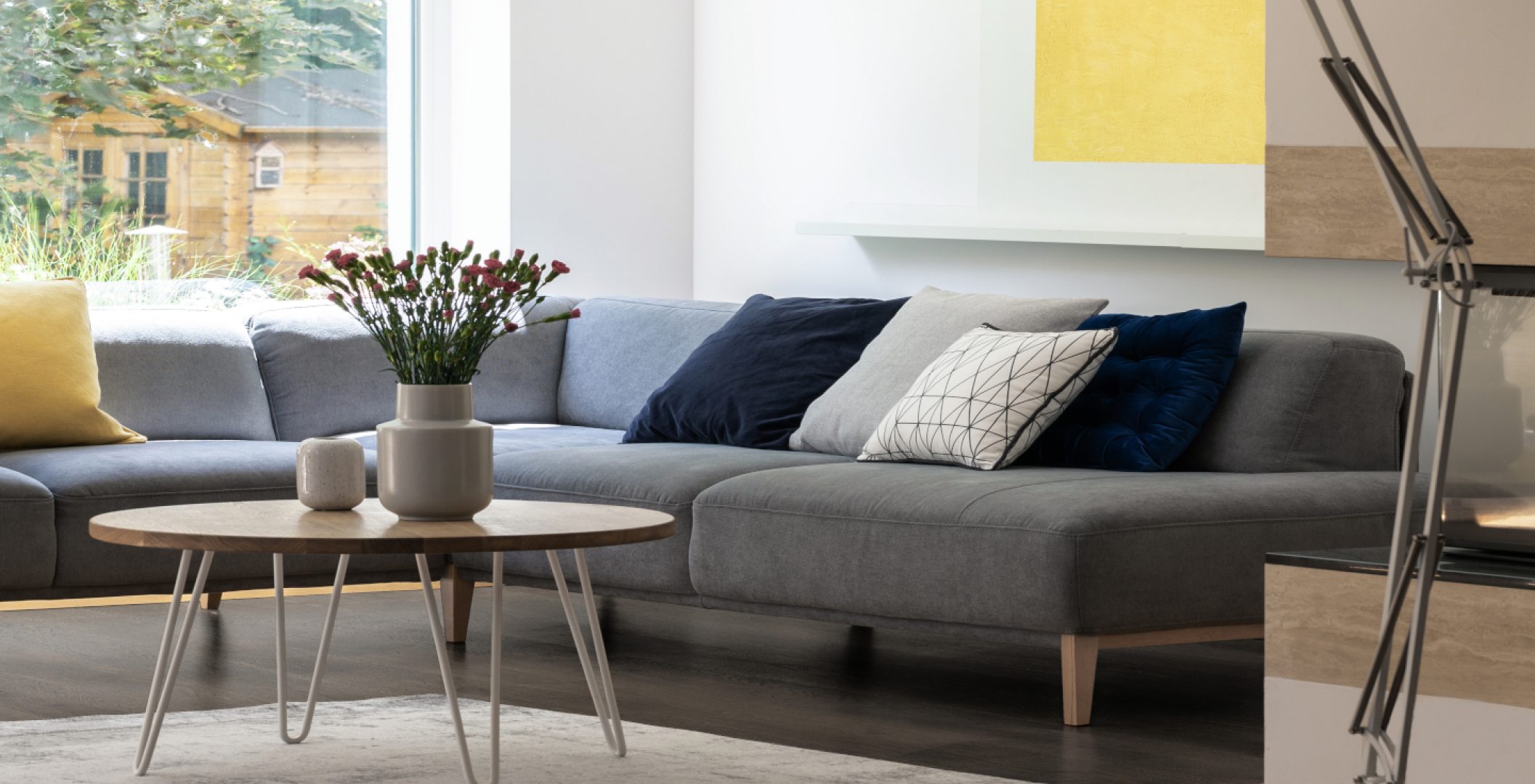 Flowers on table next to grey corner couch in bright living room interior with lamp. Real photo